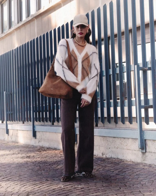 A young adult woman stands outside wearing brown pants, a white, brown and black sweater with a brown tote bag over her shoulder.