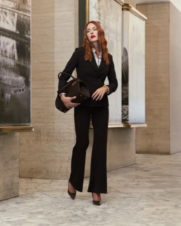 Stylish woman in a black business suit stands in a neutral-toned hallway holding a duffel bag in a fashion pose with paintings.