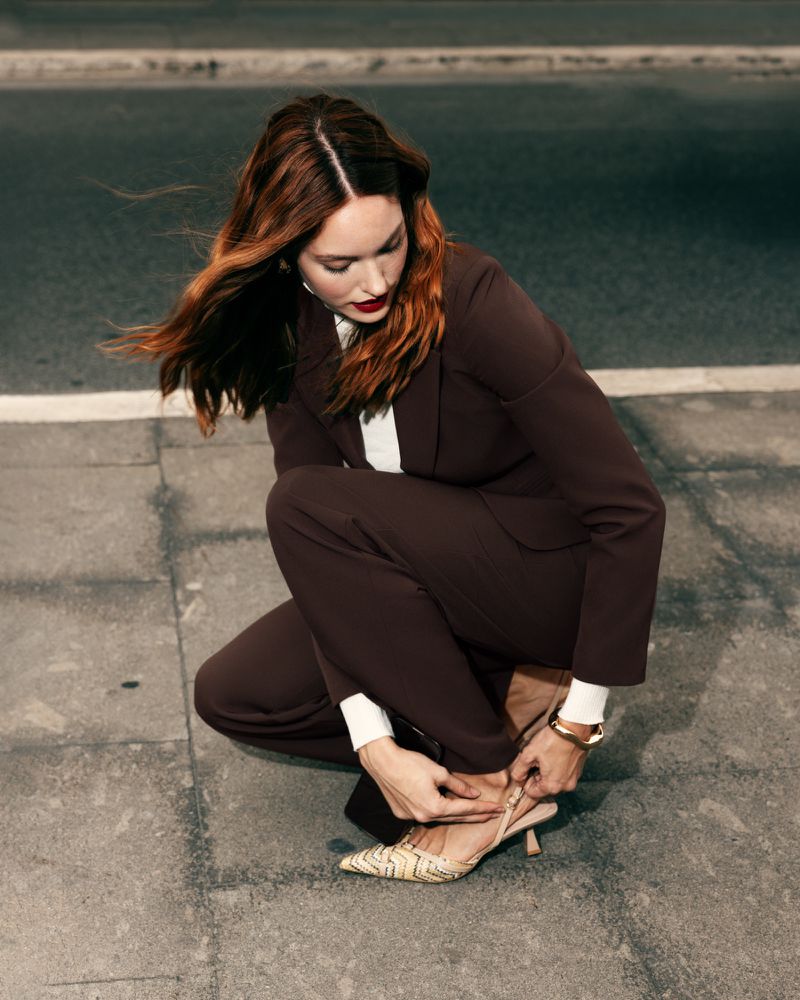 A stylish woman in a brown suit crouches on a city street, adjusting her patterned high heel shoe in an eye-level, medium shot.