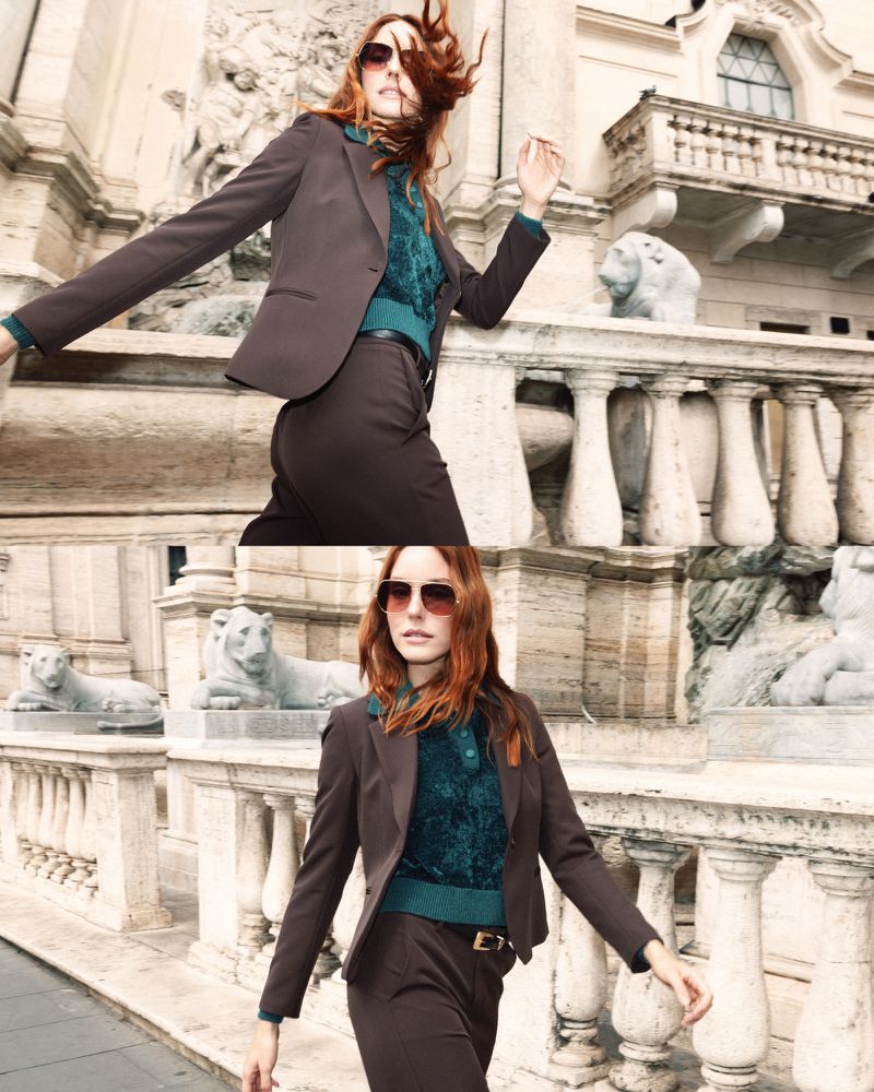A woman with red hair is wearing a brown pantsuit and sunglasses near a stone railing in an outdoor urban setting.