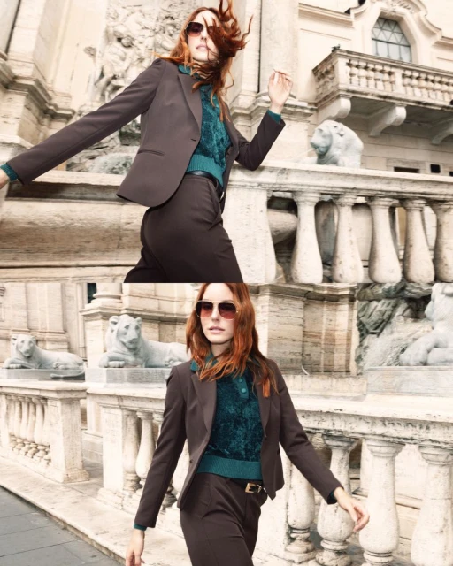 A woman with red hair is wearing a brown pantsuit and sunglasses near a stone railing in an outdoor urban setting.