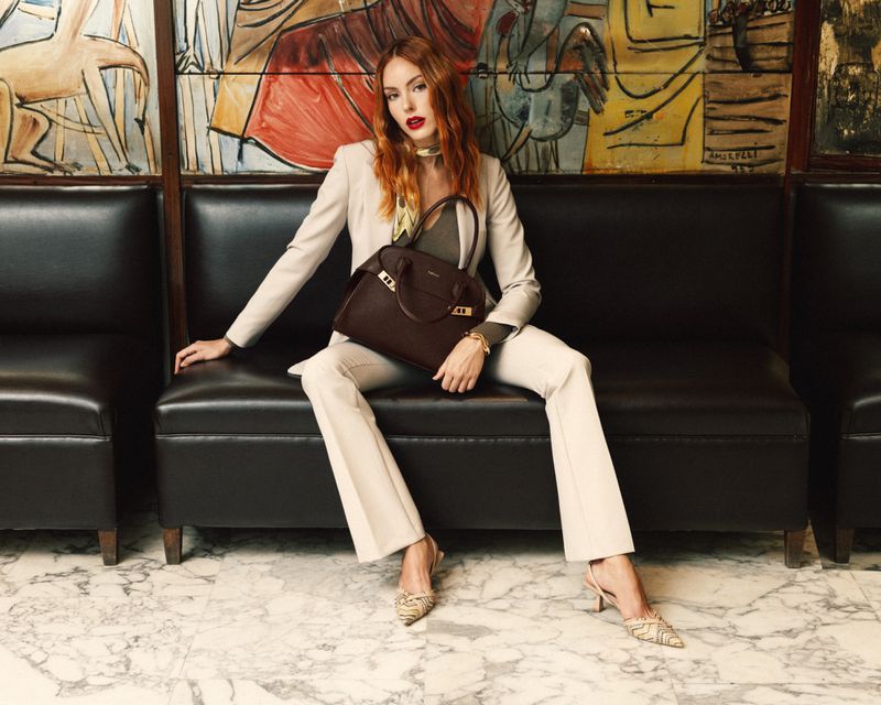A redhead woman in a beige suit sits on a black leather couch, holding a brown leather handbag, set against a colourful painting backdrop.