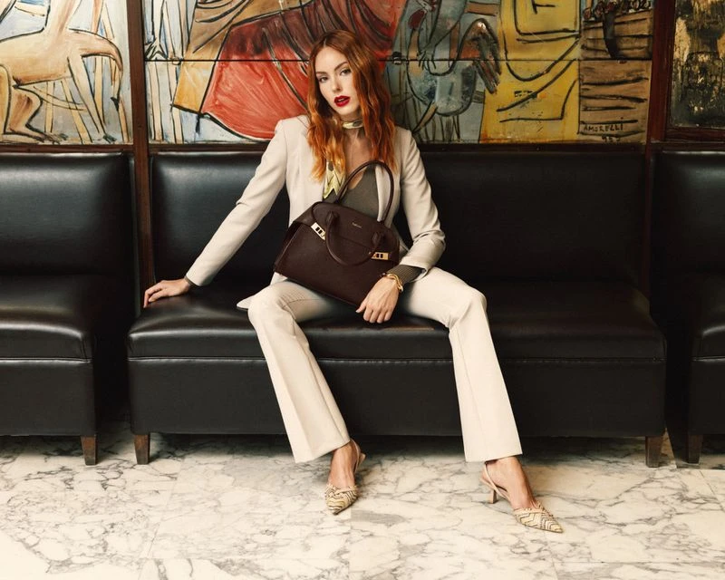 A redhead woman in a beige suit sits on a black leather couch, holding a brown leather handbag, set against a colourful painting backdrop.