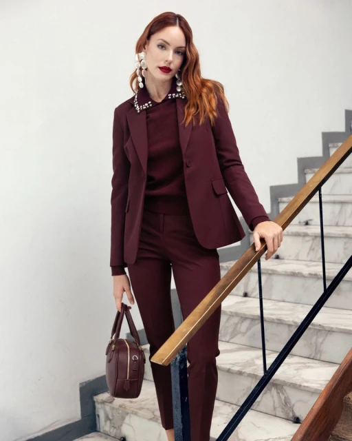 Elegant red-haired woman on marble stairs in a wine-red blazer suit with large pearl earrings and a matching handbag.