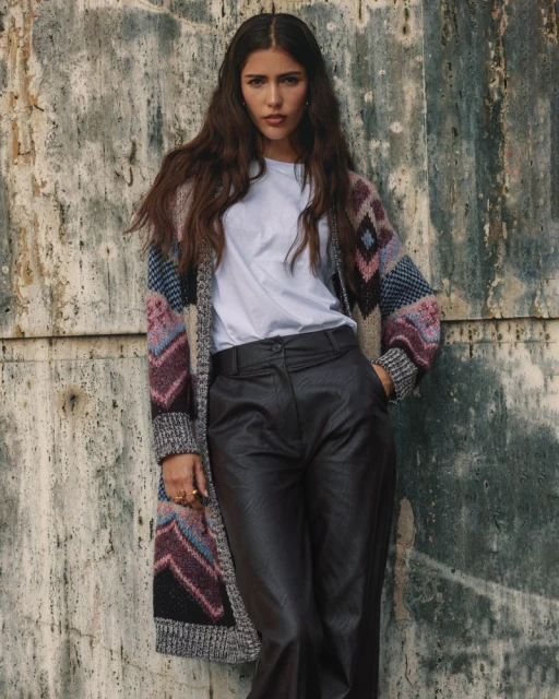 A woman with long brown hair wearing a white t-shirt, black pants, and long colorful cardigan stands against a textured wall.