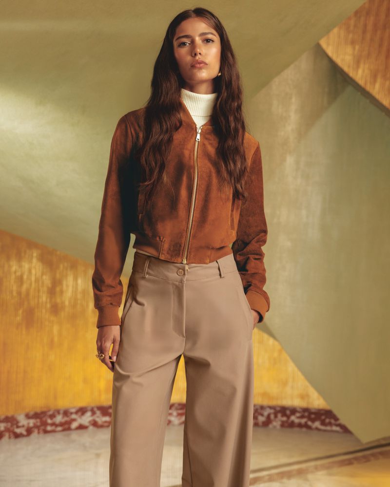 Model in brown suede bomber jacket and beige trousers against an architectural interior backdrop for Talco campaign.