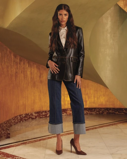 Brunette woman in a black leather jacket, white button-down, jeans, and heels poses indoors against a textured gold wall.