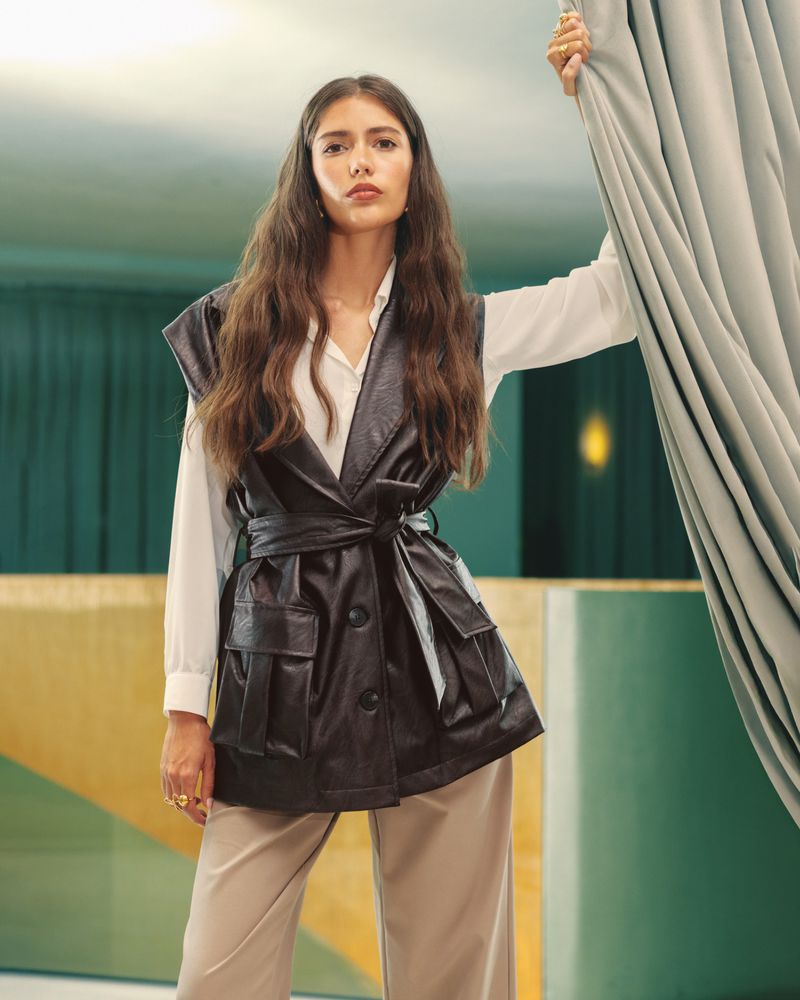 Brunette woman in a black faux leather vest and beige trousers posing in an indoor space with teal wall and a gray curtain.