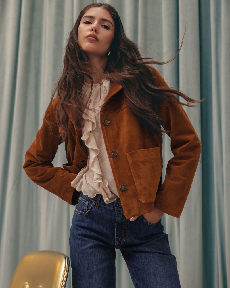 Portrait of a young adult woman posing in a brown jacket, jeans, and ruffled top, set against a light blue curtain backdrop.
