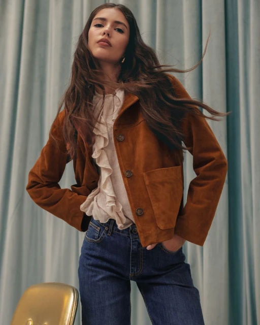 Portrait of a young adult woman posing in a brown jacket, jeans, and ruffled top, set against a light blue curtain backdrop.