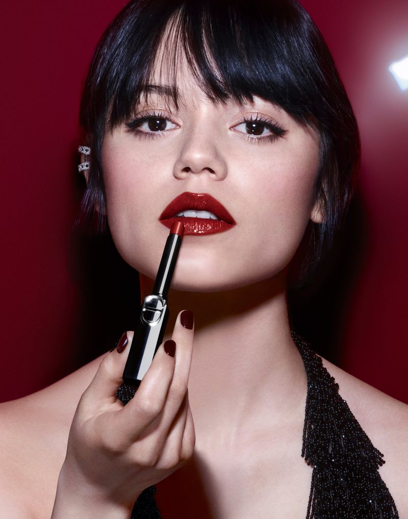 A woman with red lipstick poses in a studio setting. The subject holds a lipstick tube to her lips in a beauty shot.