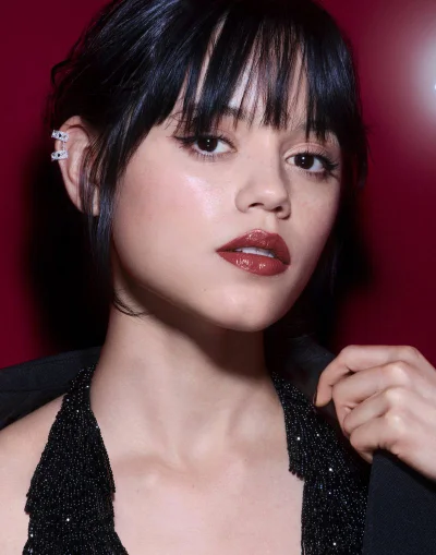Close-up portrait of Jenna Ortega with dark bangs wearing glossy deep red lipstick and a black beaded top against a rich red background.