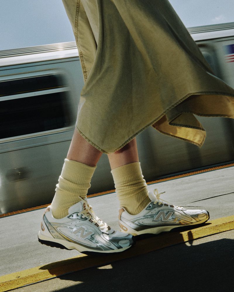 A person walks on a platform wearing New Balance sneakers, paired with beige socks and a skirt, with a train in the background.