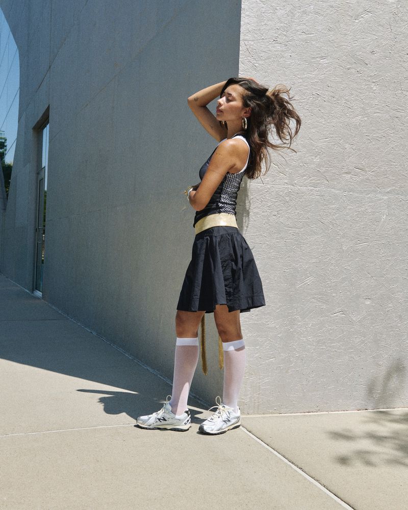 A young woman in a black skirt and white sneakers poses against a grey wall in an urban setting, bathed in bright sunlight.