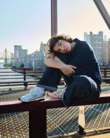 Young adult poses on a bridge wearing casual clothing and New Balance sneakers with a cityscape and blue sky in the background.