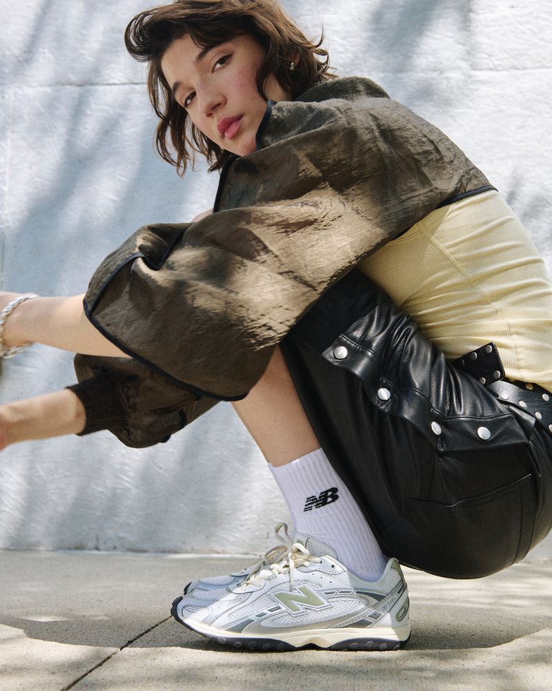 A young woman in stylish athleisurewear poses outdoors, showcasing grey New Balance sneakers with white socks and black jacket.