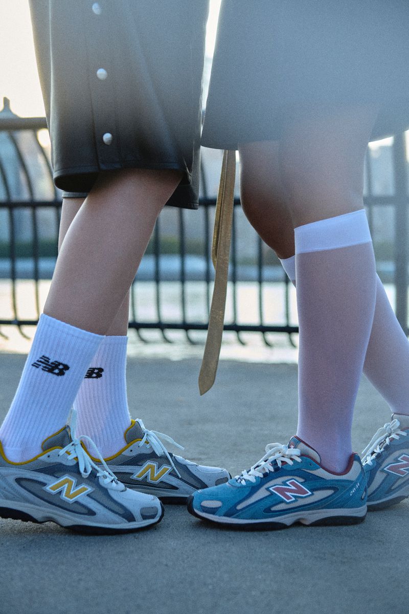 Two pairs of New Balance sneakers worn with socks are displayed outdoors against a blurred architectural backdrop, highlighting footwear fashion.