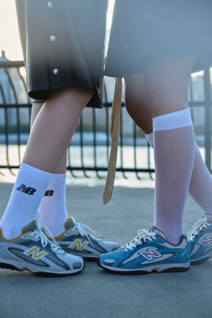 Two pairs of New Balance sneakers worn with socks are displayed outdoors against a blurred architectural backdrop, highlighting footwear fashion.