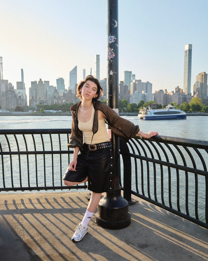 A young woman stands on a waterfront promenade next to a pole, wearing a jacket and shorts, with a city skyline and river backdrop.