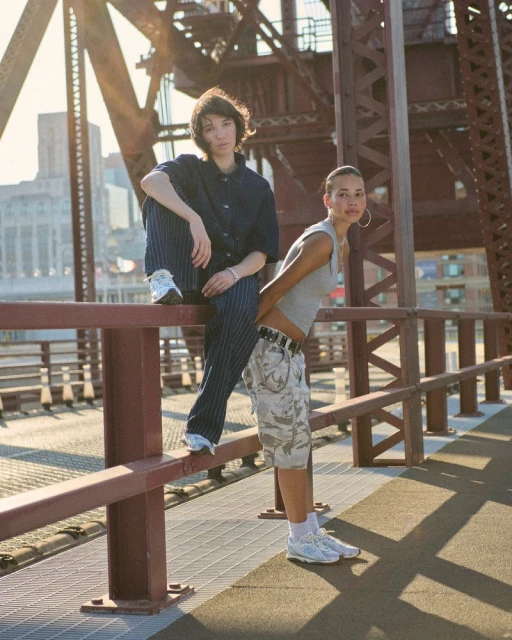 Two women pose on a bridge in a cityscape, one seated in blue pinstripes, the other in camo shorts and a grey tank top.