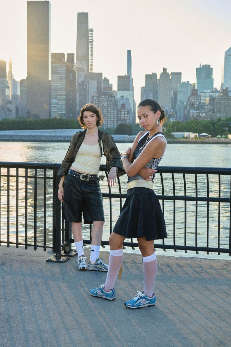 Two young women in casual streetwear pose outdoors in front of a city skyline on a sunny day, showcasing urban fashion.