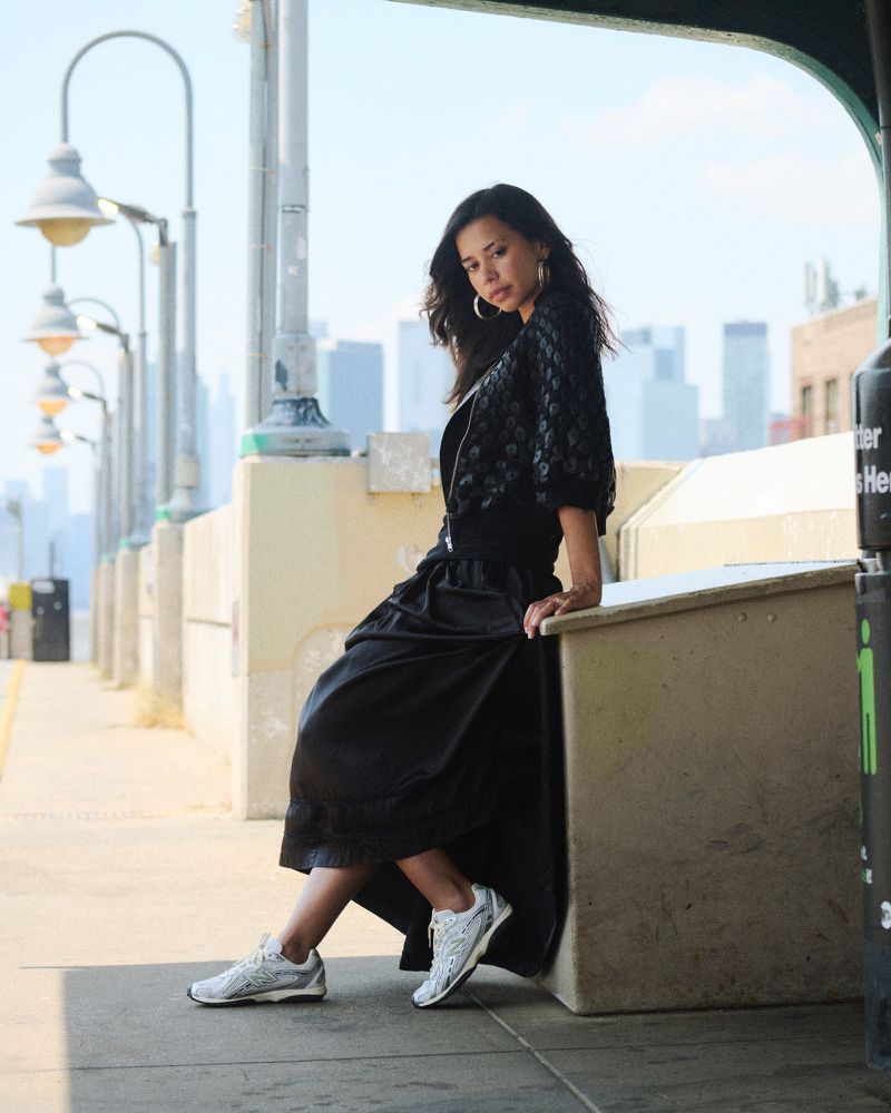 Woman in a black skirt and jacket posing on a train platform with city buildings in the background on a sunny day, street lights nearby.