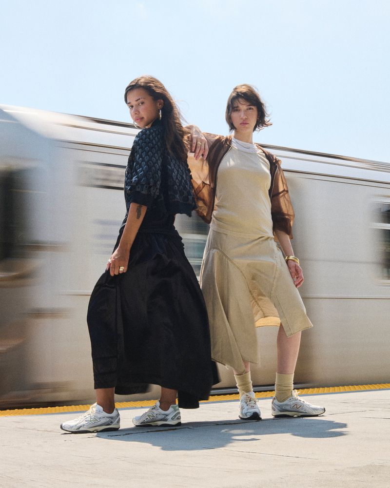 Two women pose on a train platform wearing fashionable clothing in front of a blurred train, under a bright sky.