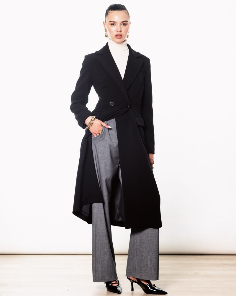 Fashionable woman in a black coat and gray pants poses indoors against a white backdrop, showcasing modern style and elegance.