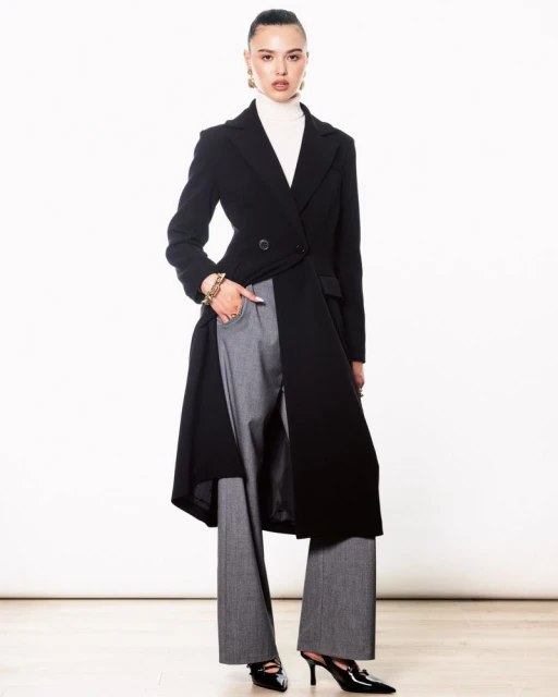 Fashionable woman in a black coat and gray pants poses indoors against a white backdrop, showcasing modern style and elegance.
