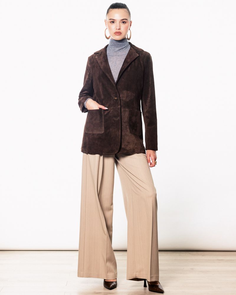 Full-length studio shot of an adult female posing in a brown suede blazer, grey turtleneck, and wide-leg beige pants with dark brown heels.