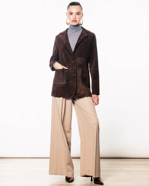 Full-length studio shot of an adult female posing in a brown suede blazer, grey turtleneck, and wide-leg beige pants with dark brown heels.