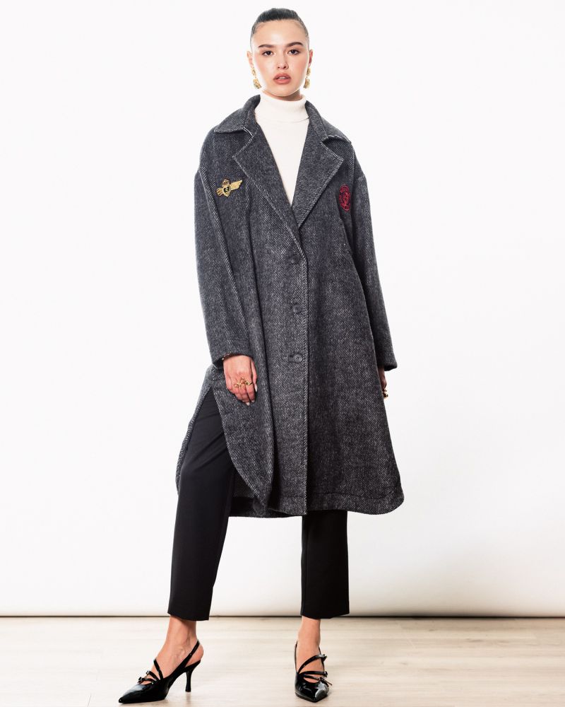 Woman in a grey wool coat, black pants, and slingback heels poses in front of a white backdrop in a fashion studio setting.