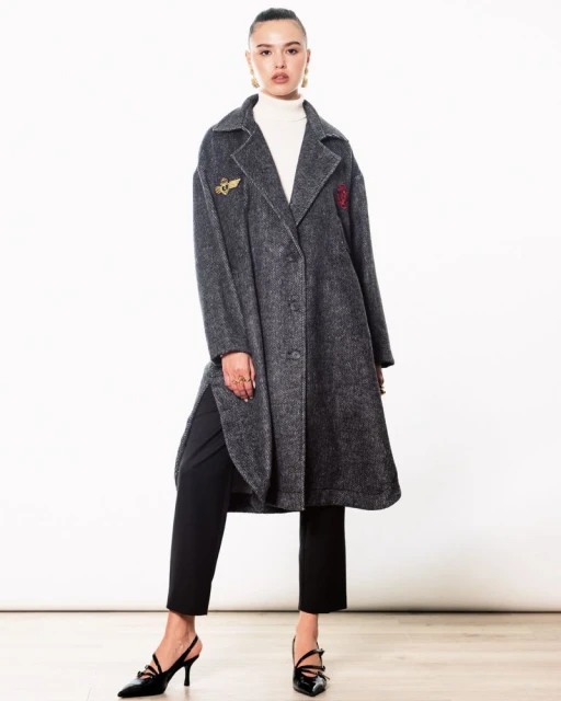 Woman in a grey wool coat, black pants, and slingback heels poses in front of a white backdrop in a fashion studio setting.
