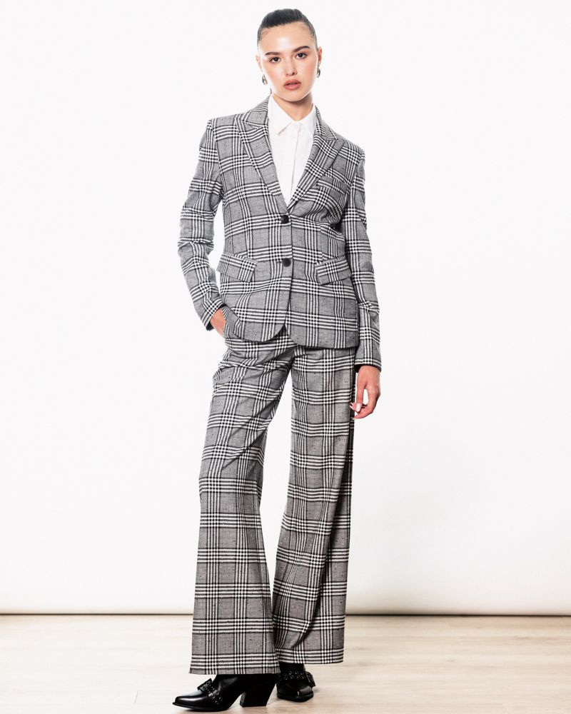 A woman in a grey plaid suit poses in a bright studio. The ensemble includes a blazer, wide-legged pants, a white button-up shirt, and black heels.