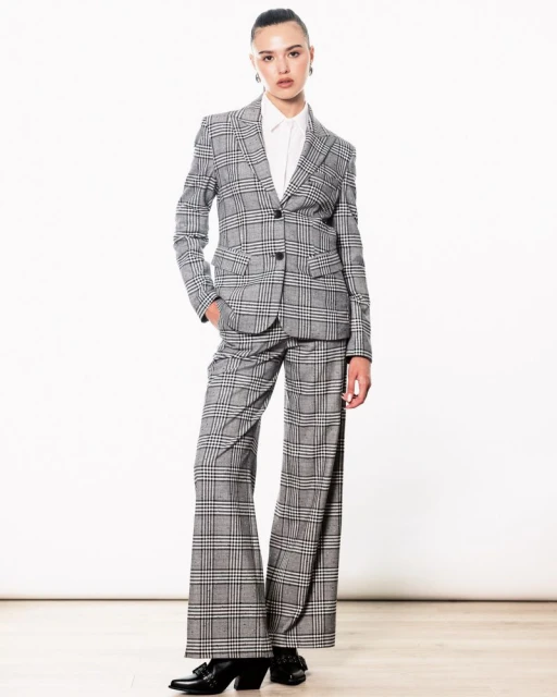 A woman in a grey plaid suit poses in a bright studio. The ensemble includes a blazer, wide-legged pants, a white button-up shirt, and black heels.