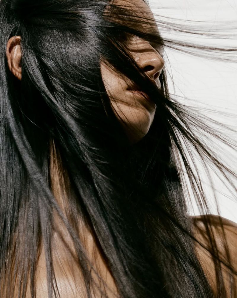 Close-up of a woman with long, dark hair blowing in the wind against a bright white background, focusing on her profile and hair texture.