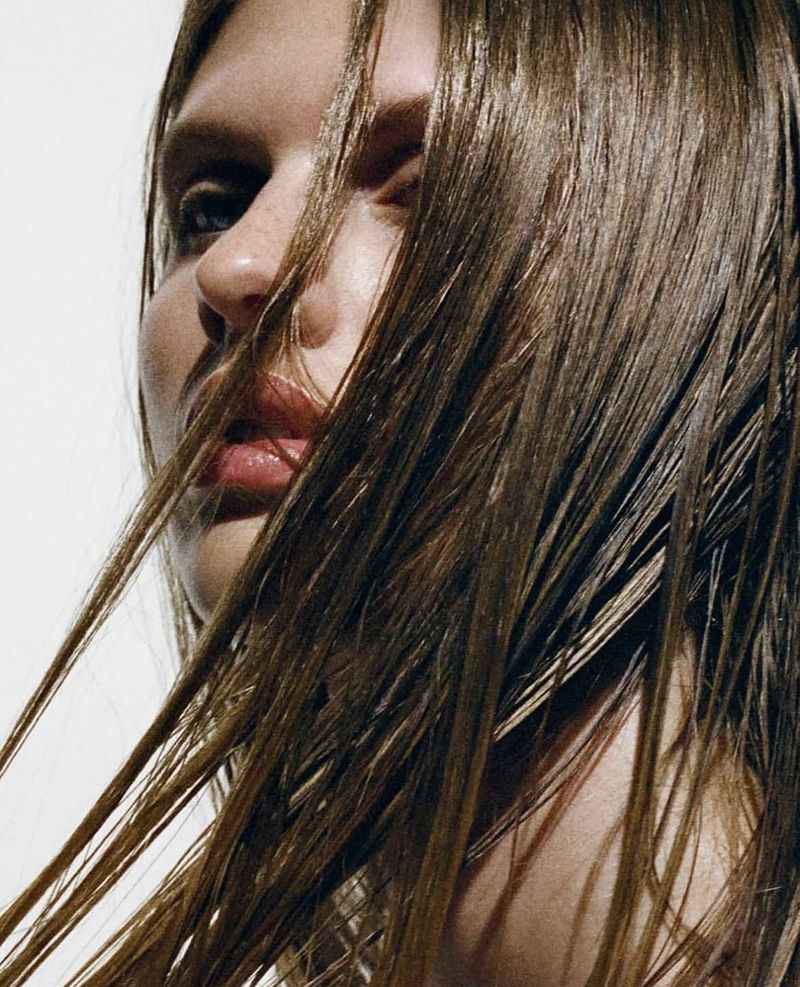 Close-up portrait of a woman with wet brown hair, neutral facial expression, and fair skin against a bright white background in a beauty context.