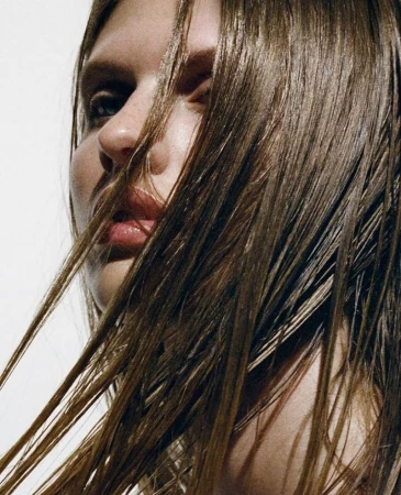 Close-up portrait of a woman with wet brown hair, neutral facial expression, and fair skin against a bright white background in a beauty context.