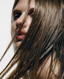Close-up portrait of a model with wet brown hair falling across her face against a white background for the Nécessaire campaign.