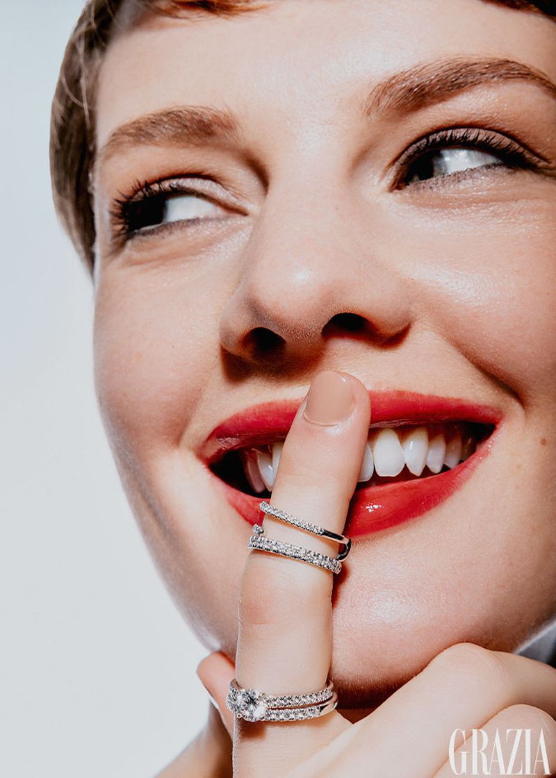 Close-up of an adult female smiling with red lipstick and diamond rings, holding a finger to her lips in a well-lit photo studio.