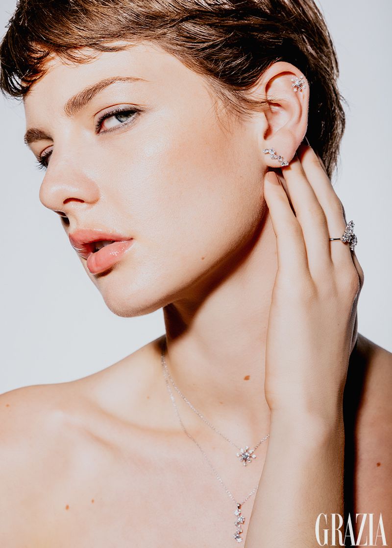 Close-up shot of a woman with short hair wearing jewelry, including earrings, a necklace, and a ring, set against a white backdrop.
