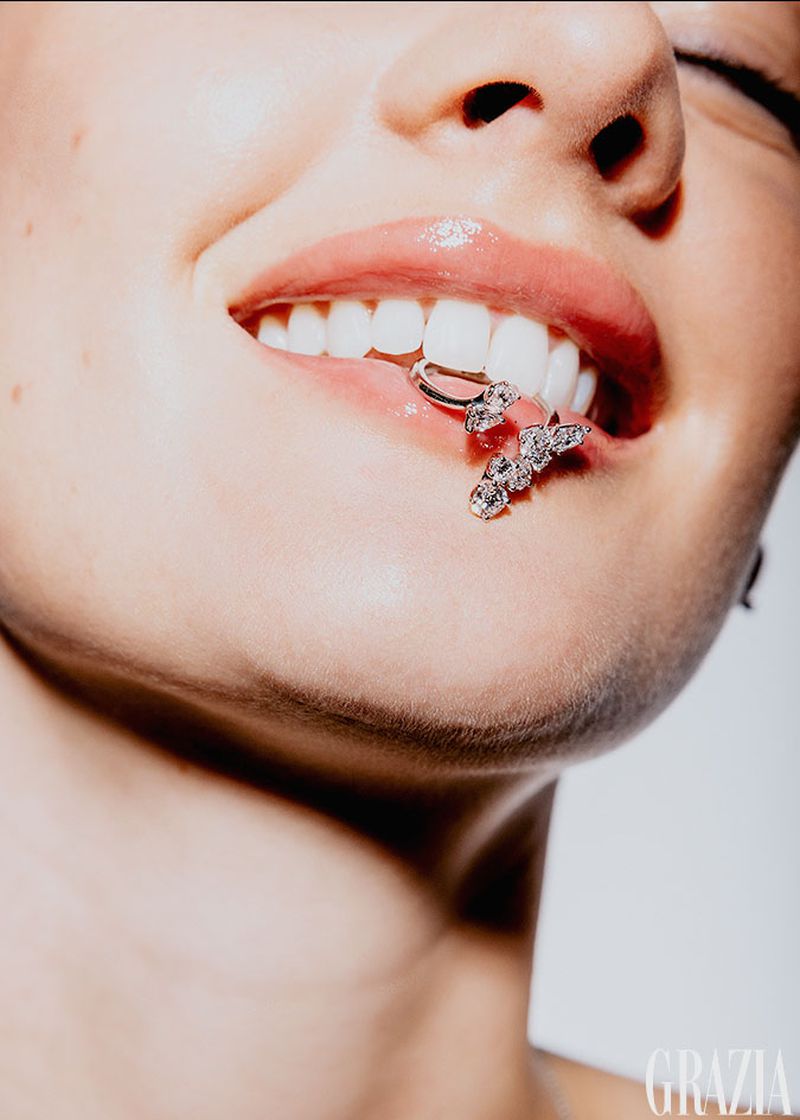 Close-up of a woman's mouth with shiny lip gloss and a sparkly dental grill against a bright white background in a studio setting.
