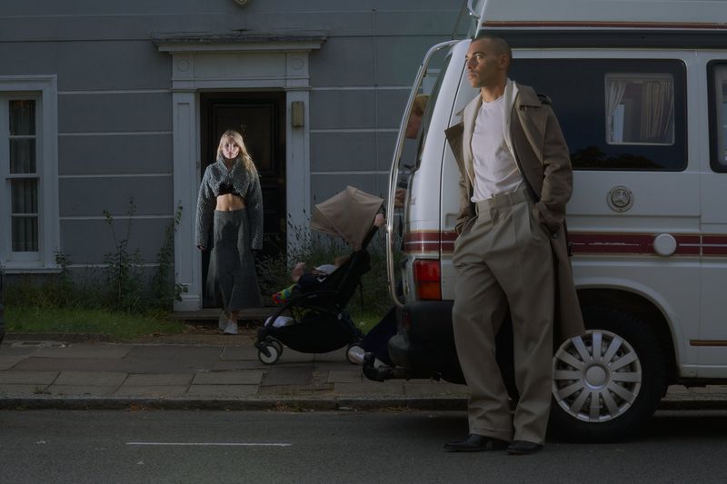 Fashion shot featuring a man in a trench coat leaning against a van and a woman in a matching outfit standing in a doorway.
