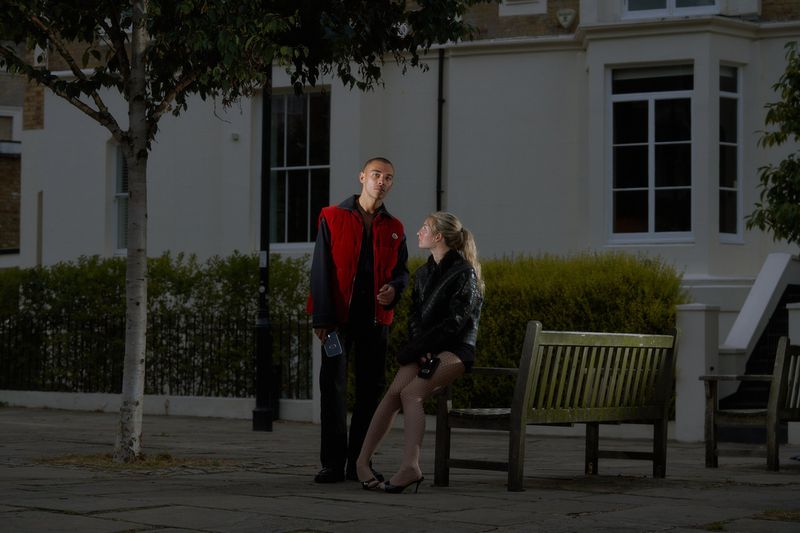 An adult man and woman converse on a city street with residential buildings; the woman sits on a park bench in the evening.