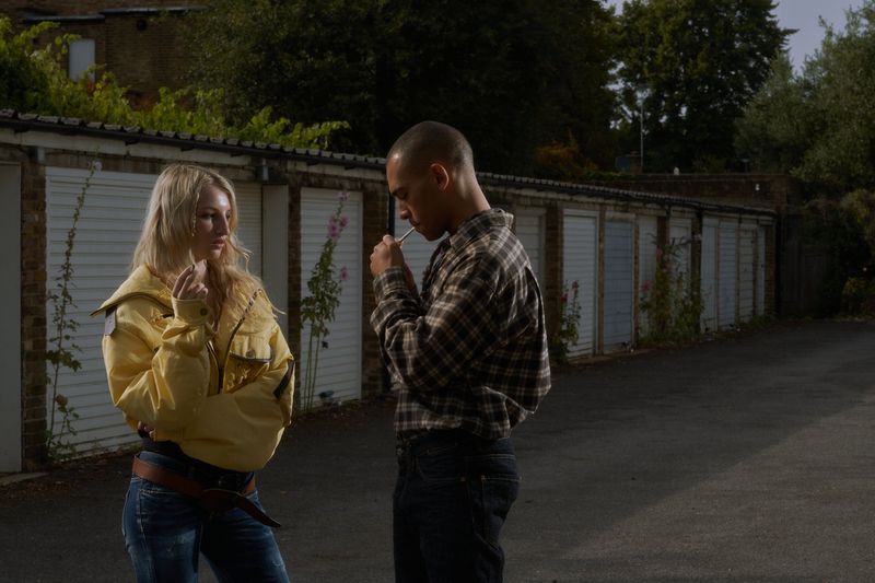 Two people are standing outside a row of garage doors, with one wearing a yellow jacket and the other in a plaid shirt smoking.
