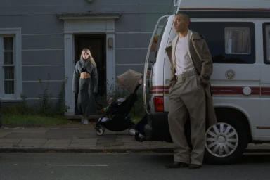 Man in a long beige coat standing beside a white camper van with a woman standing near a doorway in the background.