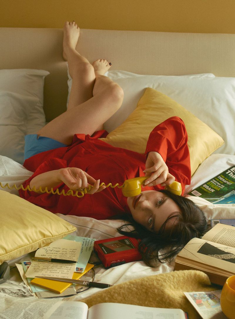 Woman in red shirt laying on bed with yellow phone, surrounded by books and papers in an indoor, casual setting.