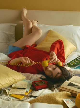 Woman in red shirt laying on bed with yellow phone, surrounded by books and papers in an indoor, casual setting.