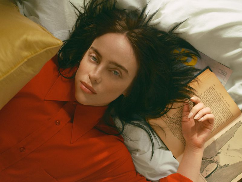 Close-up portrait of a young adult female with dark hair and light eyes, wearing a red shirt, lying on a white bed with a book.