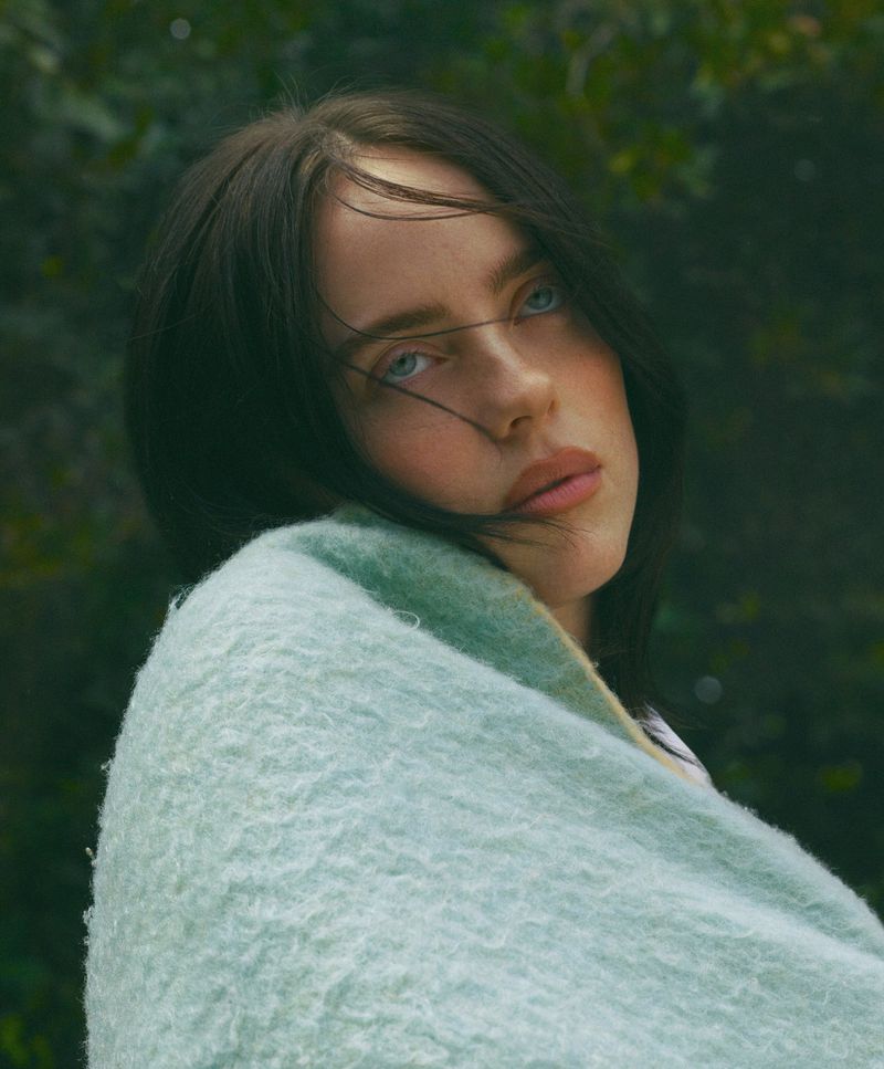 Portrait of a young fair-skinned adult woman wrapped in a pastel blue blanket, looking slightly upward against a natural green backdrop.